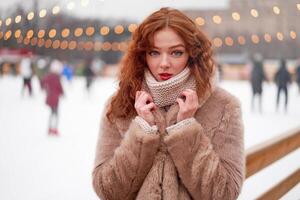 Young beautiful redhead girl freckles ice rink on background. Pretty woman curly hair portrait walking on new year fair photo