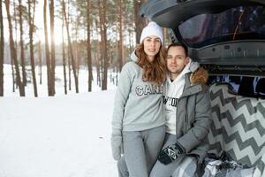 A young family of travelers guy and girl are sitting in the trunk of his car in an embrace. A walk of a young couple in the winter pine forest. Love photo