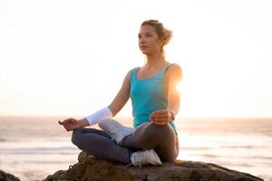 mujer práctica yoga loto actitud a meditación con verano vacaciones rocoso playa felicidad y relajación. calma hembra ejercicio con yoga meditar Oceano playa con puesta de sol dorado tiempo. foto
