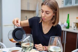 Closeup photo of women pouring tea into a cup. On the side on the table is a glass of wine and a piece of cake