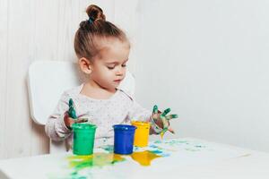 hermosa pequeño niña sorteos con dedo pinturas en un blanco sábana de papel. creativo niño desarrollo en jardín de infancia o gratis hora a hogar foto