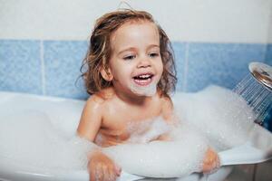 hermosa pequeño niña tomando un bañera a hogar. un linda bebé es sentado en el baño y jugando con juguetes y agua. personal higiene para niños. diario higiene foto