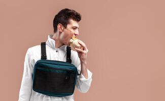 joven elegante masculino trabajador con cofre plataforma bolso es comiendo un sabroso jugoso hamburguesa en un blanco antecedentes foto