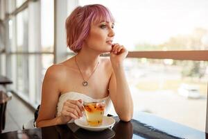 un hermosa niña con rosado pintado pelo se sienta en un restaurante cerca el ventana Bebiendo Fruta té y mirando soñando adelante. hipster niña esperando para su amigo en un fecha foto