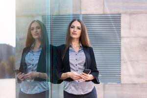 Business woman 35 years dressed stripe shirt and black jacket with long hair standing near office building outdoor use smartphone photo