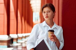 Successful businesswoman standing in central business district in modern city. Caucasian female business person with documents paper and coffee take away in white shirt photo