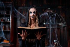 Halloween concept. Witch dressed black hood with dreadlocks standing dark dungeon room use magic book for conjuring magic spell photo