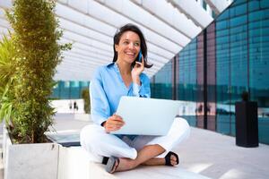 Businesswoman use laptop outdoor sitting near office building. Caucasian female business person 30 years have phone conversation and smile. Remote work freelancer concept. photo