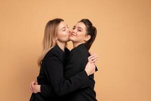 Two young business woman dressed black suit laugh and happy. Two beautiful caucasian female business person hugging isolated on beige color background photo