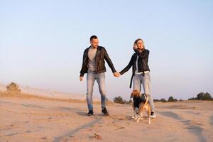 Young beautiful caucasian couple wearing leather jacket and jeans walks desert sand with Beagle dog best friend. photo