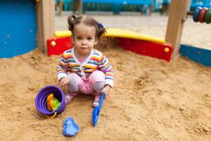 a little girl with two tails is dressed in a striped colorful jacket is playing in the sandbox on the playground photo