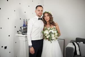 reunión el novia y novio en el dormitorio, el recién casados son contento. hombre vestido en el wgite camisa y corbata de moño mujer vestido en el blanco Boda vestir y guirnalda peinado con ramo de flores en mano foto