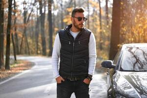 A handsome young Caucasian man wearing style black vest standing near black car on nature. photo