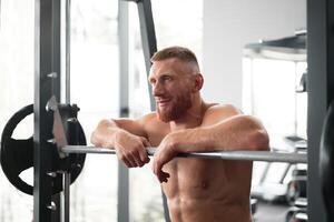 Gym man leaning barbell resting after hard training. Handsome caucasian athlete take break. photo