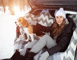 un joven familia de viajeros mujer son sentado en el maletero de su coche en un abrazo con su amigo perro beagle. invierno bosque en el antecedentes. de viaje con un perro por coche foto