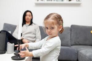 Little girl stands near the sofa at home, pulls his hands to the candlestick, looking into the camera. On the side, her mother sits and watches the girl photo