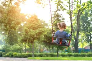 Unrecognizable Little Caucasian girl riding swing at playground sunny summer day rear view photo