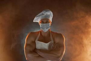 nuevo normal concepto. muscular cocinero vistiendo protector médico mascarilla, posando con doblada brazos en ahumado antecedentes foto