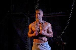 Young shirtless muscular man standing among metal chains, looking at camera, copy space photo