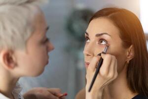 maquillaje artista aplicando brillante base color sombra en del modelo ojo y participación un cáscara con sombra en antecedentes foto