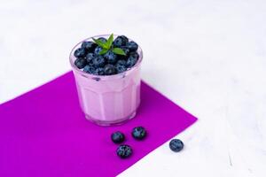 Tasty fresh blueberry yoghurt shake dessert in glass standing on white table purple napkin background. photo