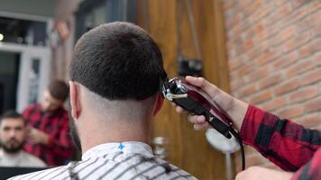 jovem à moda cabeleireiro dentro uma xadrez vermelho e Preto camisa faz uma corte de cabelo em a costas do a do cliente cabeça dentro uma barbearia video