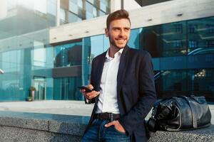 Millennial businessman with a mobile phone in his hands. Young successful business stylish man with a black leather bag walks on a city street against the background of office buildings photo