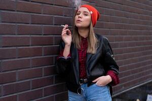 Beautiful young stylishly dressed Caucasian girl smokes cigarette on the street. photo