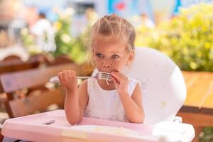 linda pequeño caucásico niña comiendo espaguetis a mesa sentado en niño asiento al aire libre restaurante. foto