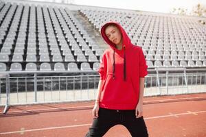 un hermosa joven niña de mezclado carrera soportes en un Deportes estadio corriendo pista vestido en un rojo capucha con un capucha. ropa de deporte para formación en el otoño en frío clima foto