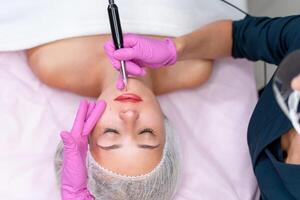 Cosmetology cabinet client lies on couch. Beautician applies marking on lips. photo