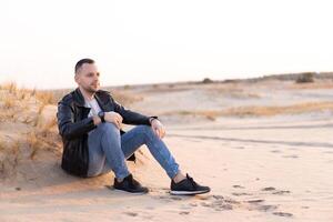 joven caucásico hombre vestido negro cuero chaqueta y azul pantalones se sienta en arenoso playa . hermoso europeo chico camina solo foto