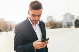 milenario empresario mira dentro el pantalla de su móvil teléfono. de cerca retrato. joven exitoso, elegante negocio hombre camina en un ciudad calle, acecho social redes en su teléfono inteligente foto