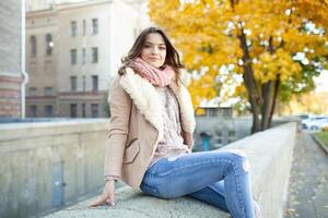 Beautiful caucasian brunette girl sitting warm autumn day with background of trees with yellow foliage and a city. photo