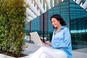 Self employed woman working with laptop sitting outdoor near corporative office building. Pretty business woman freelancer do remote work sitting on bench in downtown smile. Positive business person photo