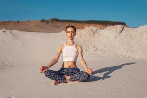 A beautiful young Caucasian girl in a white top and wide pants sits in a lotus position on the beach on the sand. photo