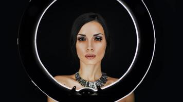 A beautiful young Caucasian girl with a beautiful make-up looks out from behind a circular lamp for portrait photo shooting