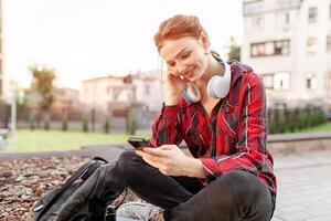 A red-haired girl with freckles is dressed in a red checked shirt is sitting in a lotus pose in the park listening to music on headphones surprised looks at the phone screen. Good news. Modern student photo