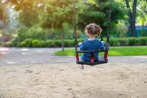 Unrecognizable Little Caucasian girl riding swing at playground sunny summer day rear view photo