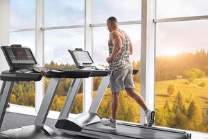 Muscular athletic bodybuilder fitness model running treadmill gym near big window photo