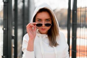 al aire libre cerca arriba retrato de joven hermosa mujer con largo pelo en Gafas de sol, vestido en un blanco suéter, cerca el campo deportivo foto