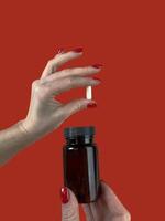 Woman holding pill and bottle on red background, closeup. Space for text photo