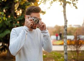 Young handsome hipster guy walks in a beautiful autumn park on the background of yellow leaves in warm sunny weather and takes pictures on a pig film camera. Autumn leisure time. Creative youth photo