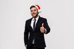 joven hermoso caucásico chico en negocio traje y Papa Noel sombreros soportes en blanco antecedentes en estudio carita y demostración pulgares arriba. foto