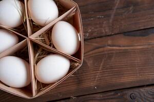 White chicken eggs lie in round wooden basket which stands on a dark wooden table. photo