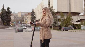 jong elegant blond meisje in een beige loopgraaf jas en jeans staat met een elektrisch scooter vith de straat Aan de achtergrond video