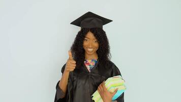 A young African-American woman in a black robe and a master's hat stands straight holding textbooks and shows a good gesture. She looks at the camera. Portrait of half height video