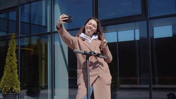 A young student girl in a sweater, jeans and a coat with an electric scooter stands near a stylish modern office center building and takes a selfie. Front view. Ecological city transport video