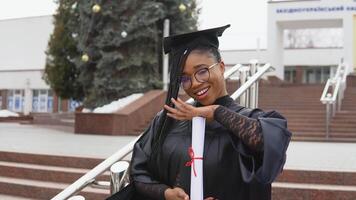 A young woman at the university at the master's mantle holds a diploma and looks at the camera. Portrait with the stairs in front of the central entrance to the university on background video