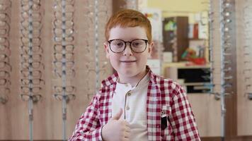 en rödhårig pojke i en pläd röd och vit skjorta står med transparent glasögon för syn korrektion och visar en tecken med hans finger upp ser hetero in i de kamera video